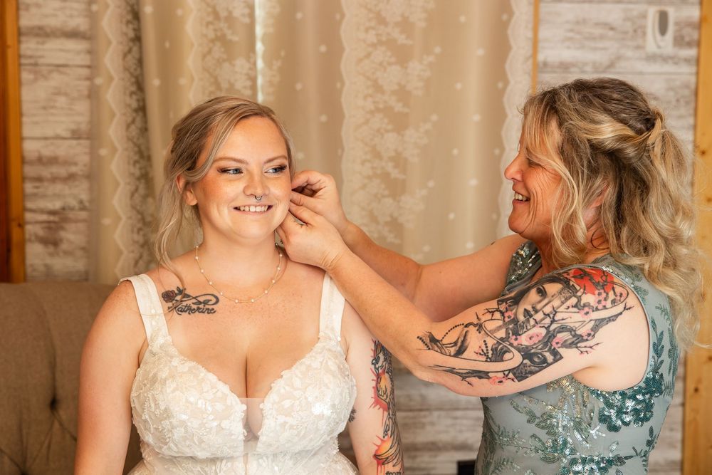 Bride in lace wedding dress smiles as tattooed bridesmaid helps put on earrings in a warmly lit room.