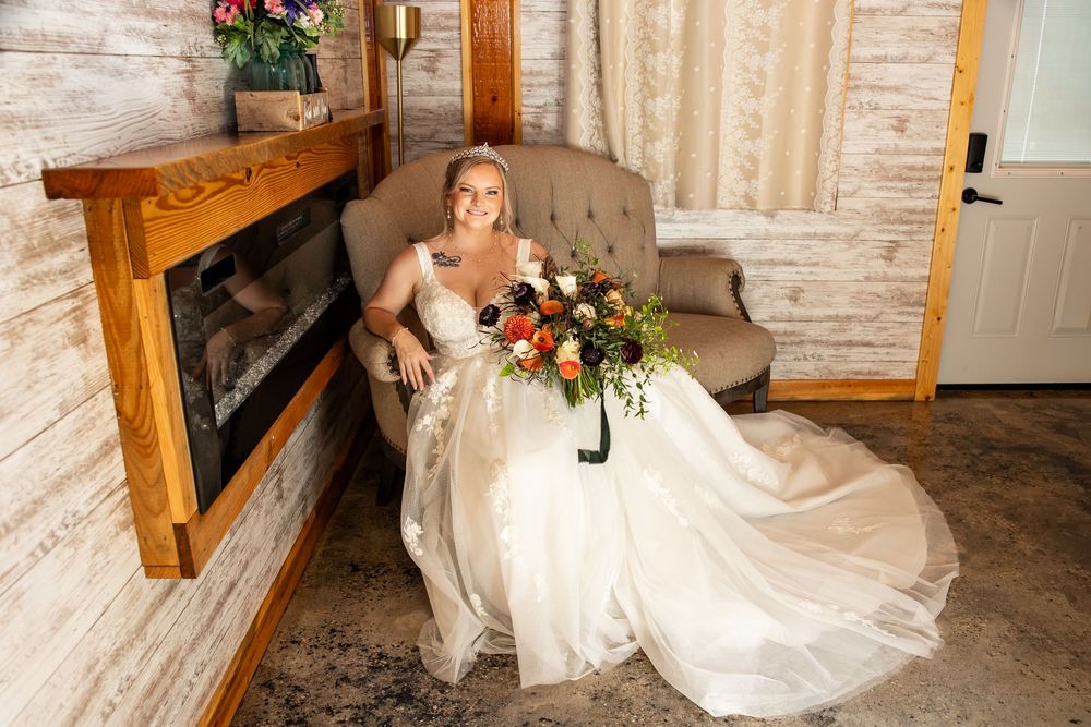 Bride in white gown holding colorful bouquet sits elegantly beside a rustic fireplace in a cozy cabin setting.
