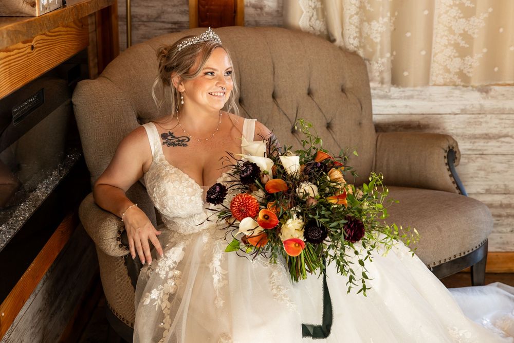 Bride in lace gown holding colorful fall bouquet seated in tufted chair near fireplace, wearing tiara and smiling.
