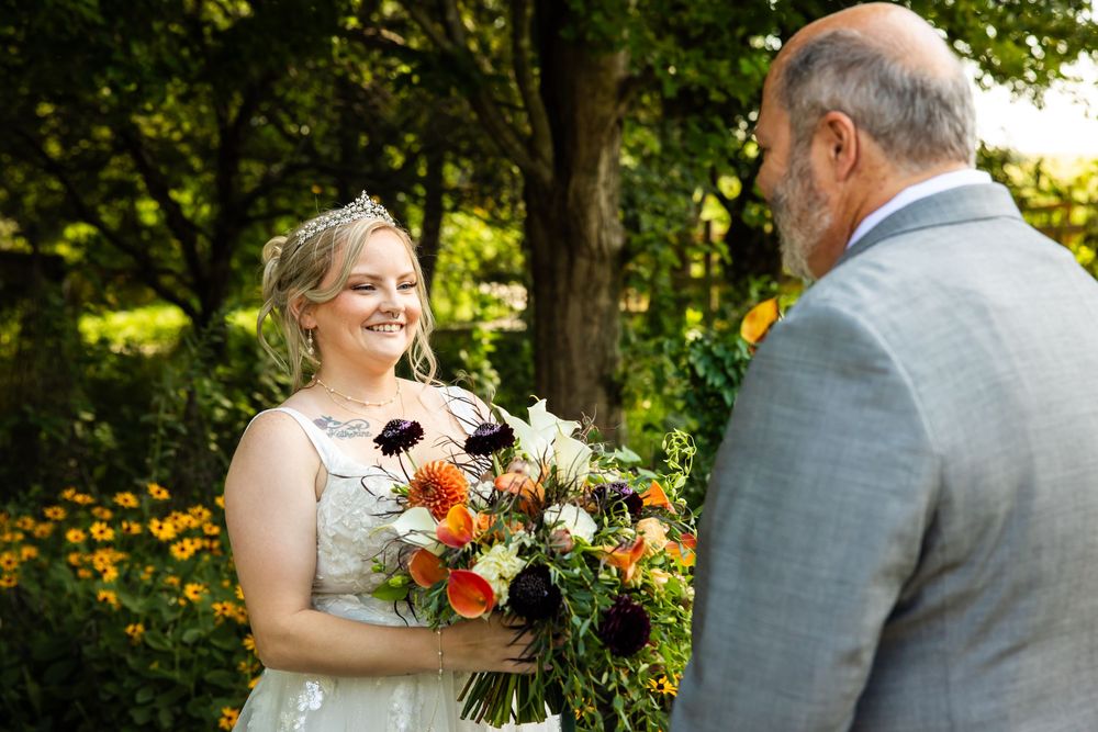 Bride holding colorful bouquet of orange and white flowers smiles at older man in gray suit outdoors.