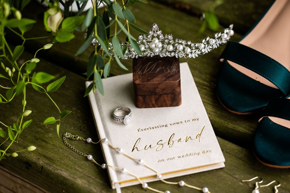 Wedding flat lay with pearl tiara, vow book, diamond ring, teal heels, and greenery on rustic green surface.
