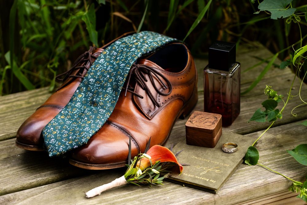 Brown leather Oxford shoes with floral tie, boutonniere, cologne, and ring on rustic wooden surface.