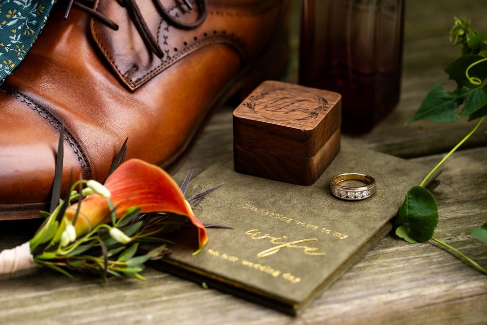 Wedding flat lay with brown leather dress shoe, wooden ring box, gold band, calla lily bouquet, and wife journal on rustic wood.