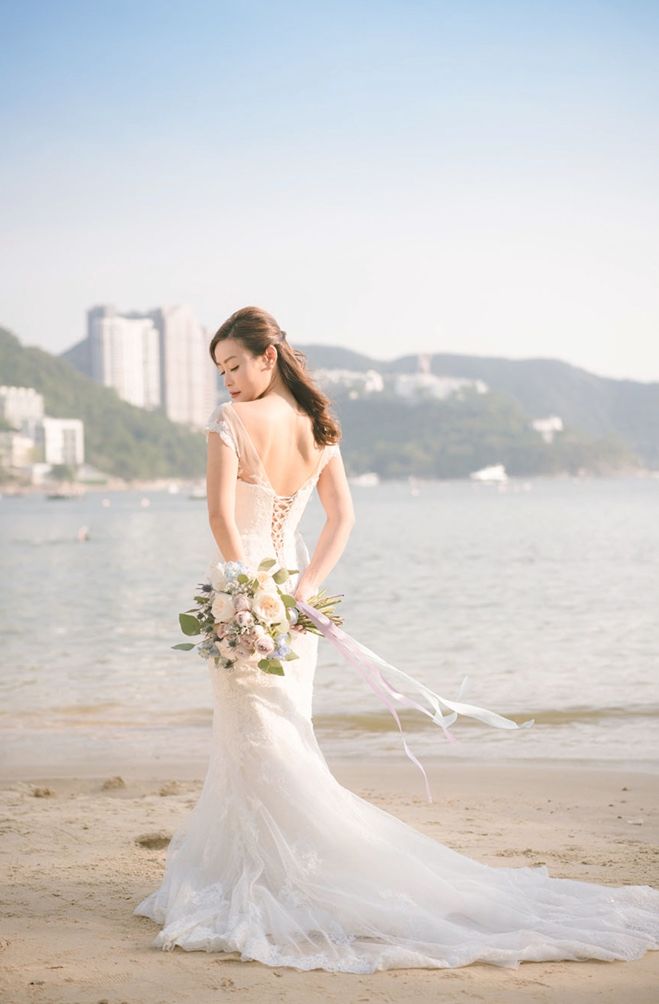 A bride in an elegant lace wedding gown with corset back details poses with bouquet on a misty beach at sunset.