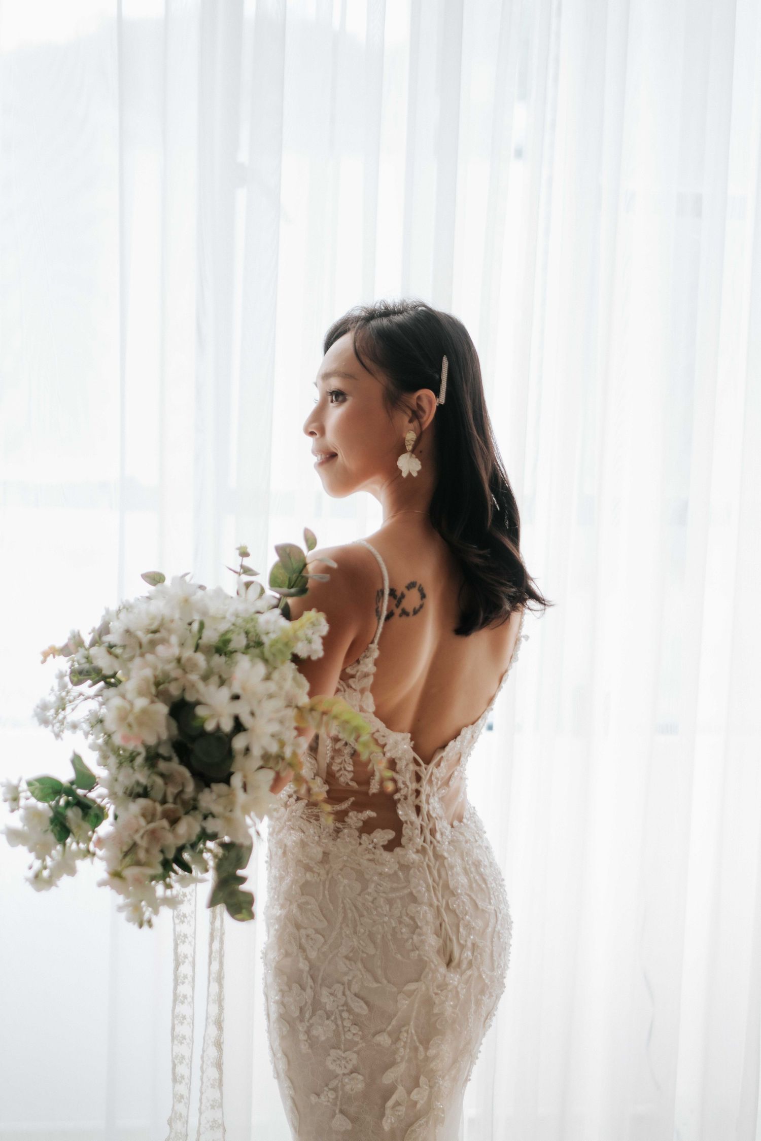 A couple embraces intimately by a sheer white curtained window during a romantic wedding day photoshoot.