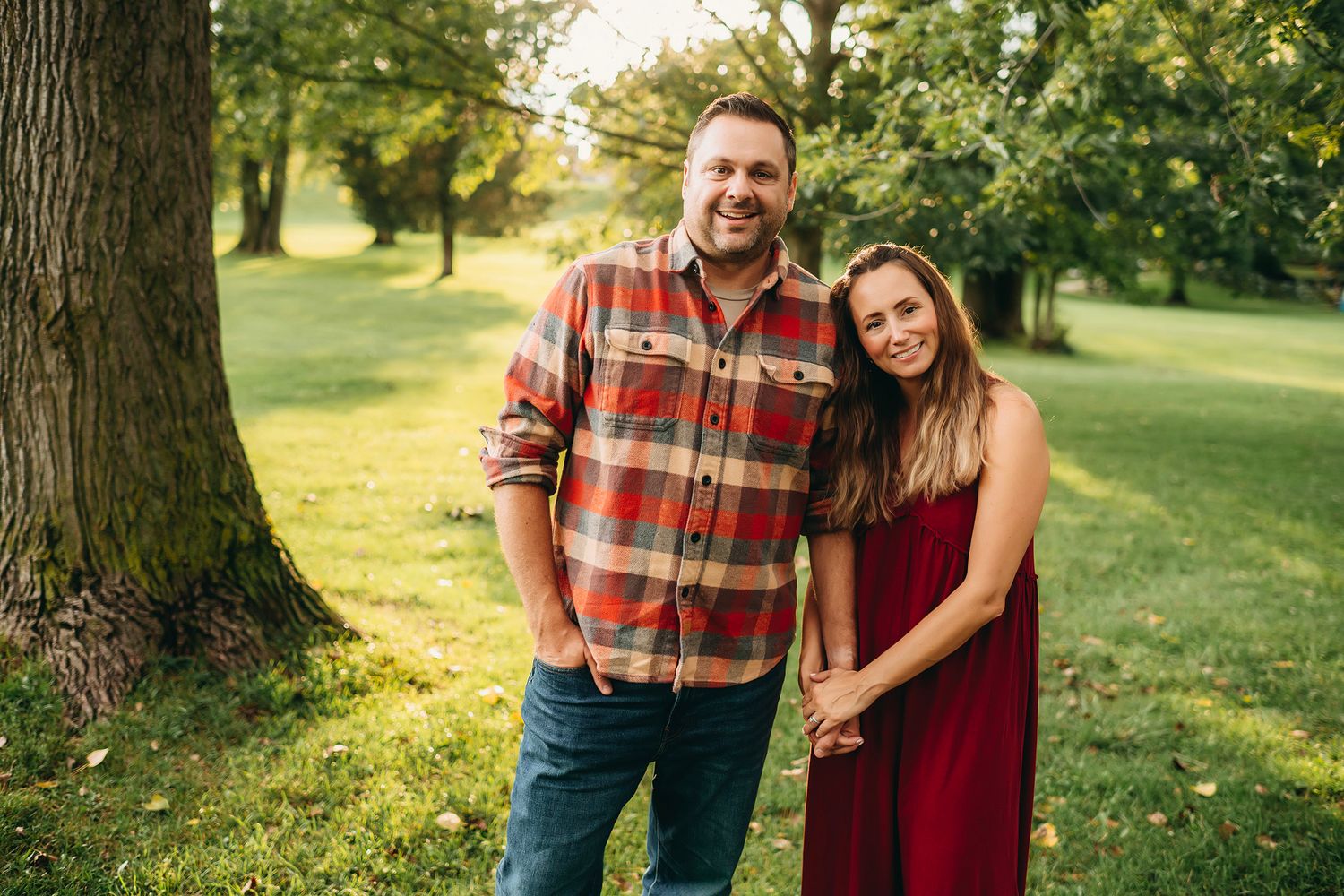 A smiling couple holds hands in a lush green park, man in plaid shirt and woman in red dress.