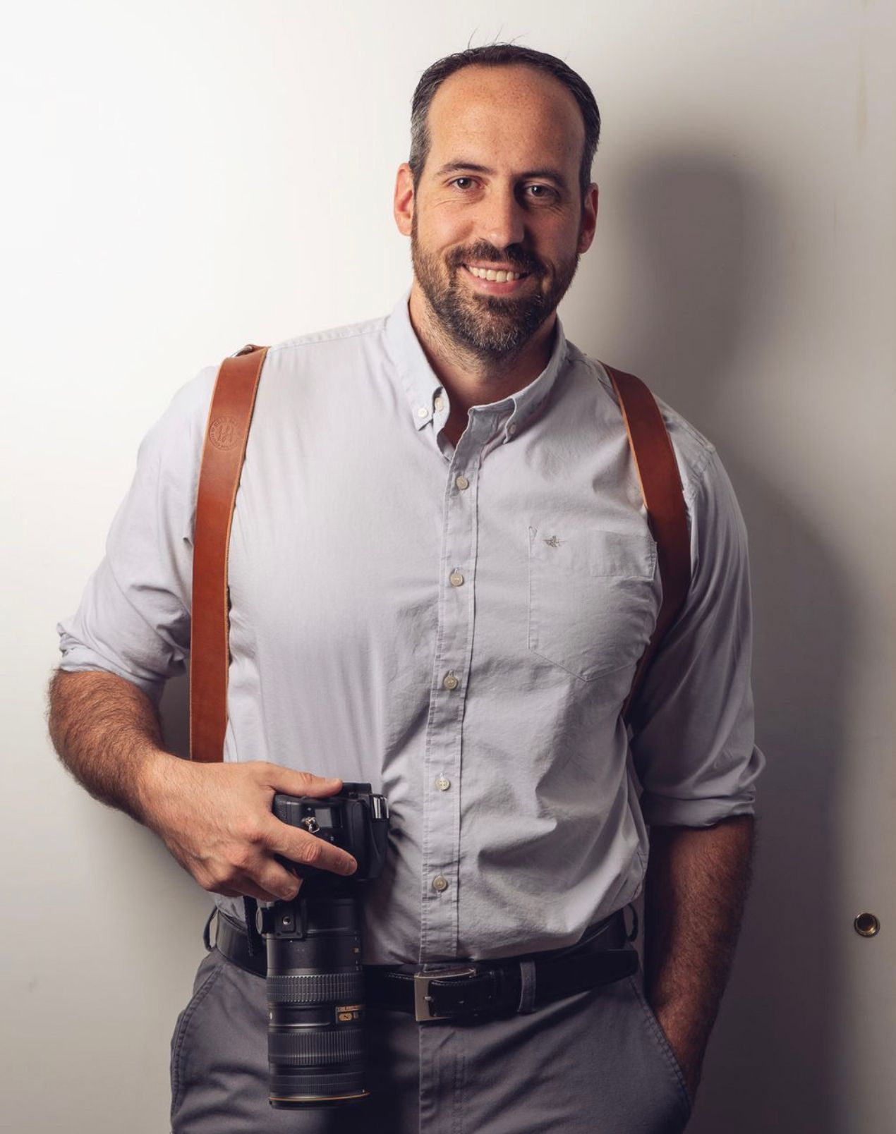 Smiling man with beard holding DSLR camera with brown leather strap, wearing light blue shirt and gray pants.