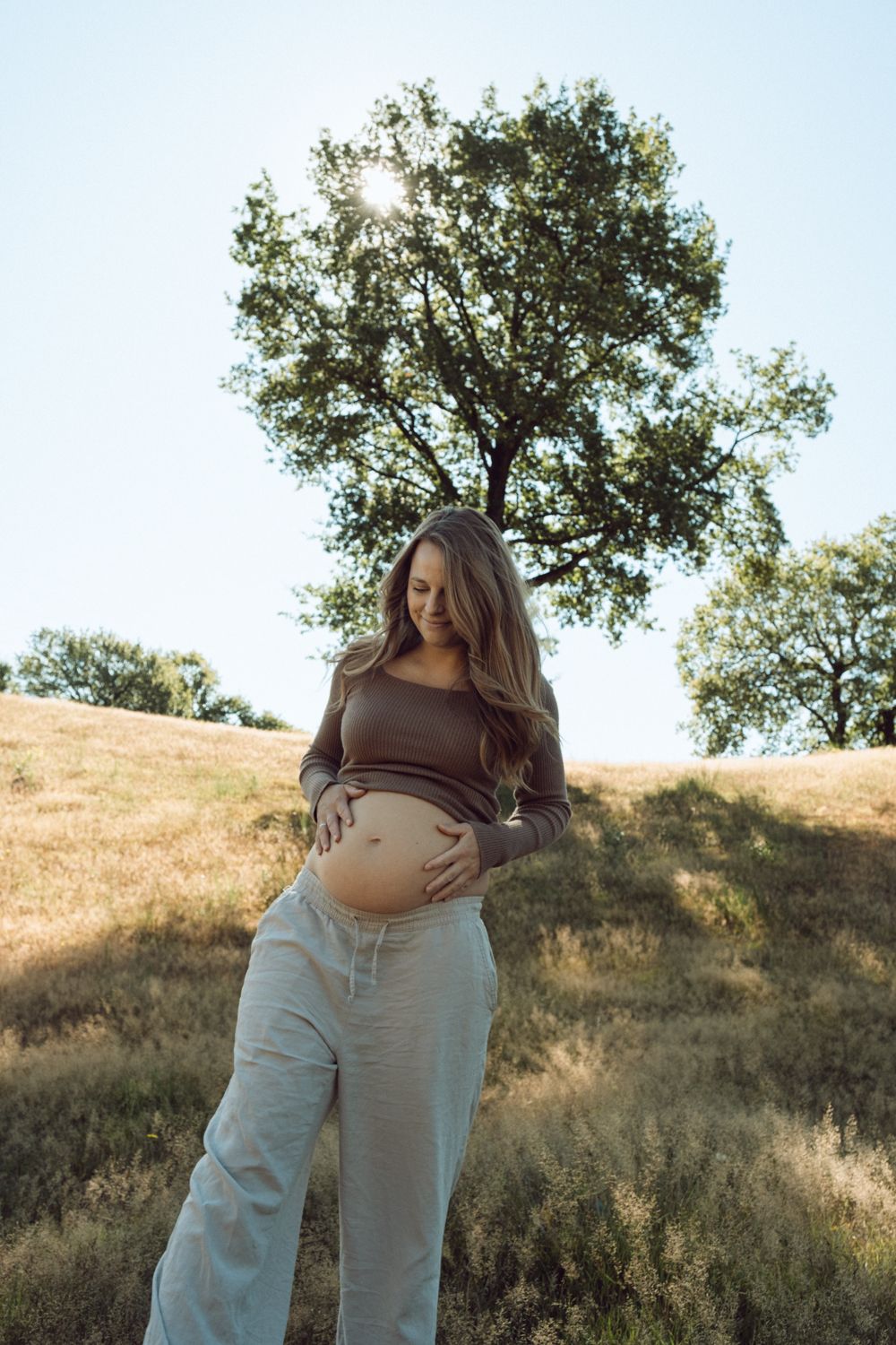 Smiling pregnant woman in brown top and gray pants holds her baby bump outdoors in a sunlit grassy field.