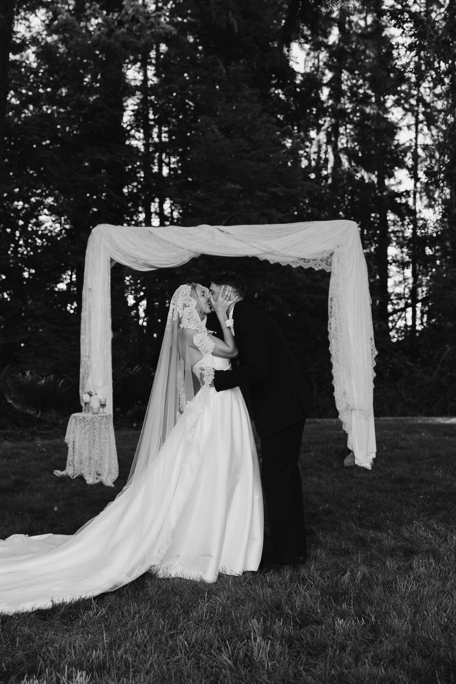Bride and groom sharing a kiss beneath a flowing lace wedding veil in golden sunlight outdoors.