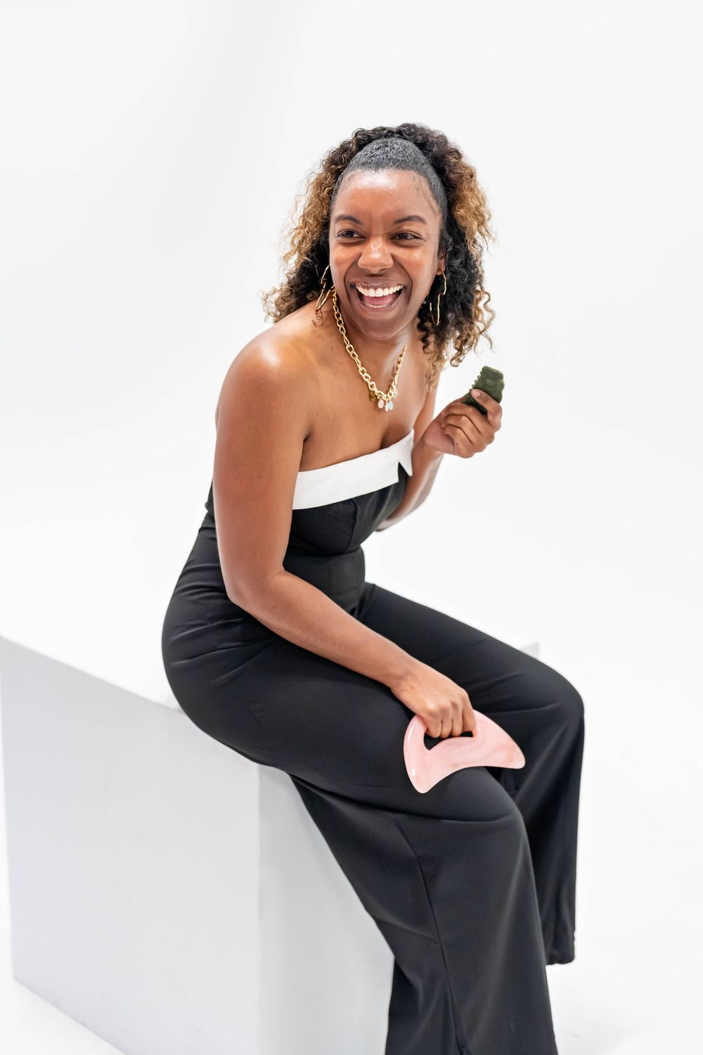 Smiling woman in black strapless jumpsuit sitting, holding jade roller and gua sha beauty tools on white background.