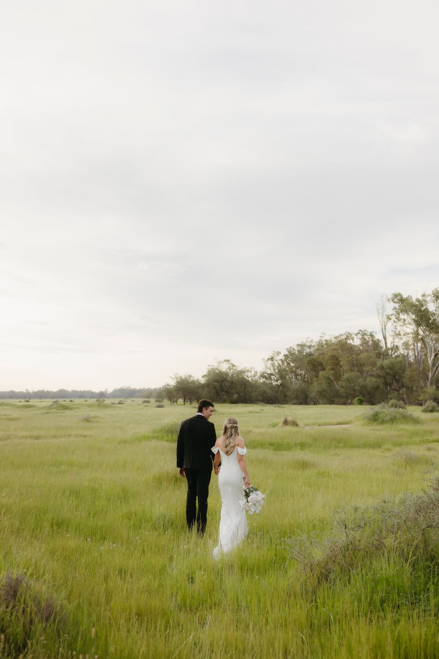 Bride and groom walking hand in hand through a vast green meadow on their wedding day.