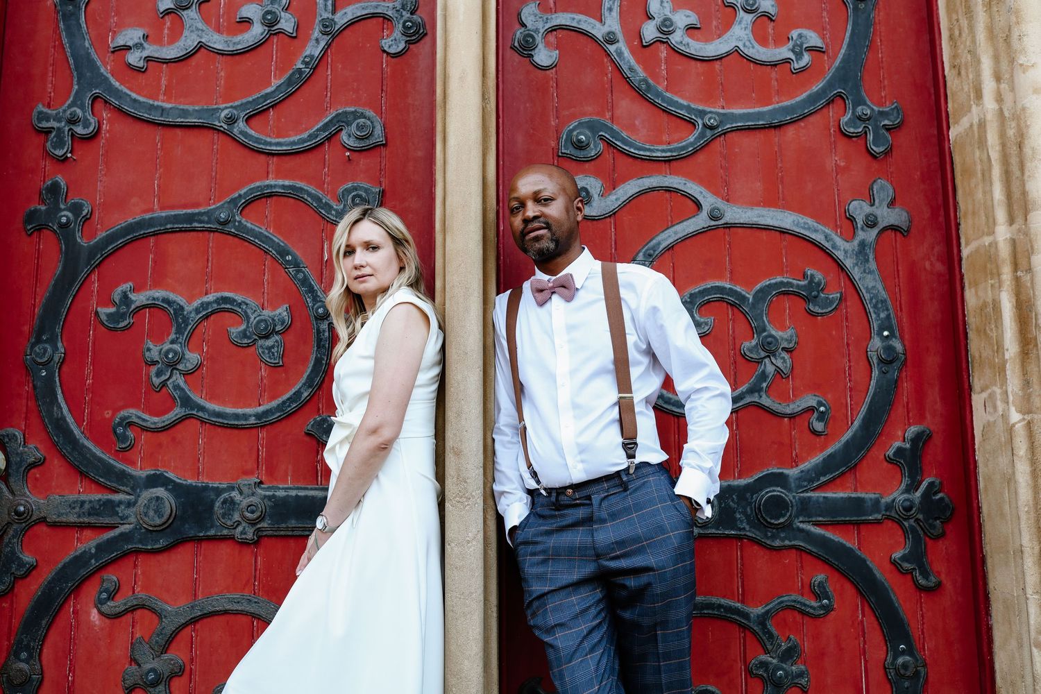 Elegant interracial couple posing before ornate red doors, bride in white gown, groom in suspenders and bow tie.