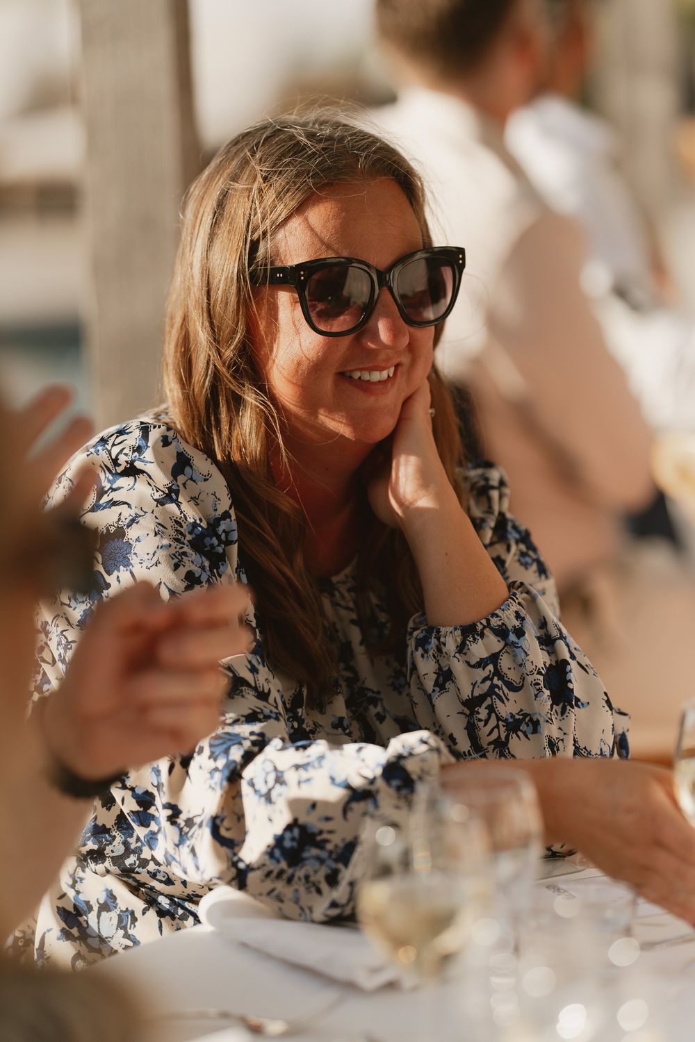 Smiling woman in floral dress and sunglasses enjoying outdoor dining with a glass of white wine.