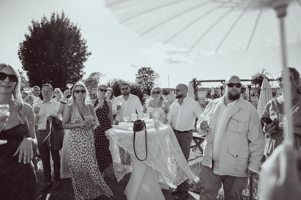 Black and white photo of elegantly dressed guests mingling outdoors at a summer wedding reception under a white parasol.