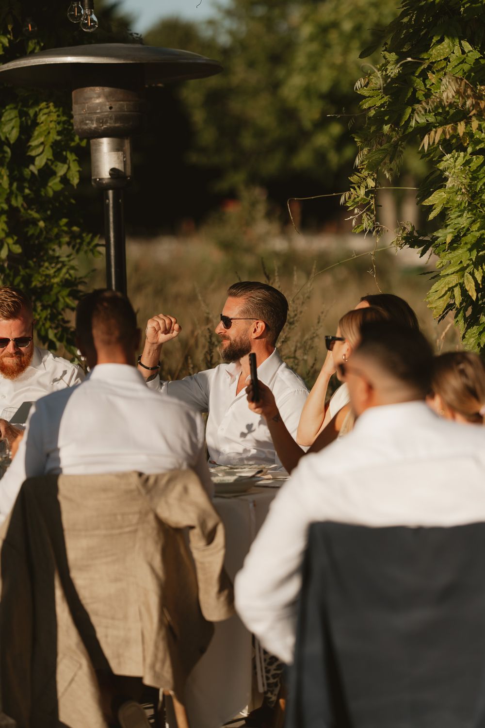 Guests in white attire celebrate outdoors at a sunny garden party near a patio heater surrounded by lush greenery.