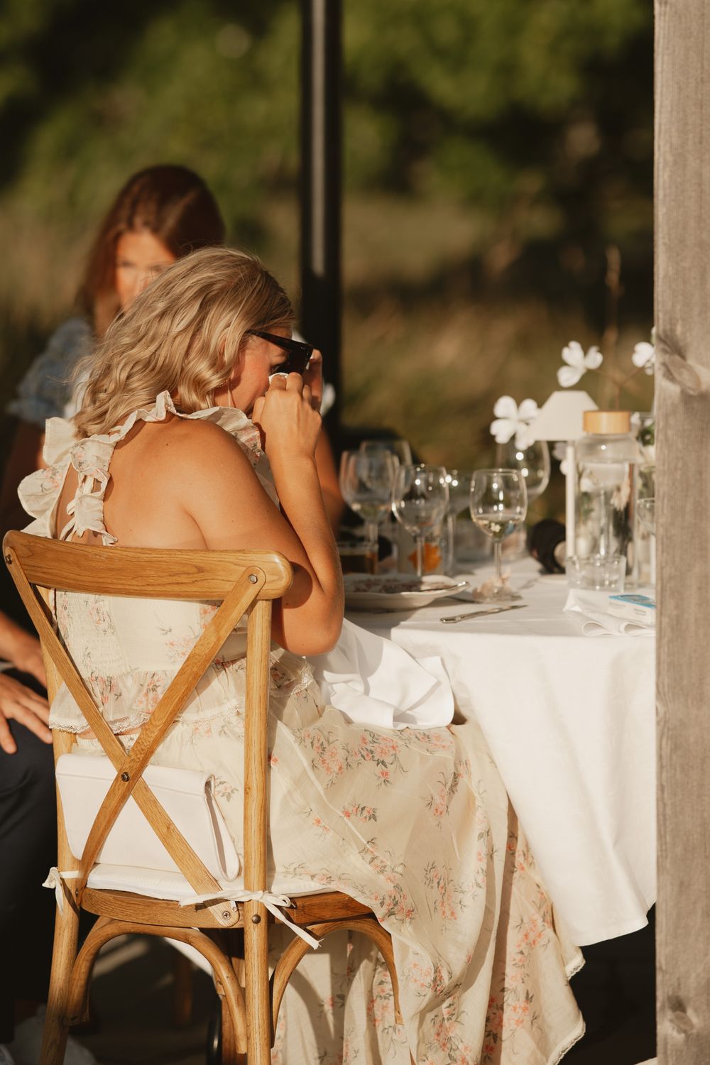 Blonde woman in floral dress sitting at elegant outdoor dining table, wiping tears at a wedding reception.