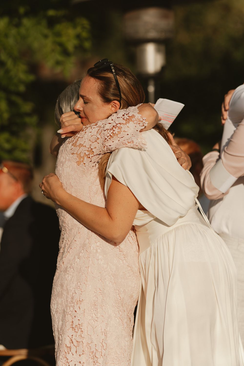 Two women in elegant dresses share an emotional hug at an outdoor wedding ceremony.