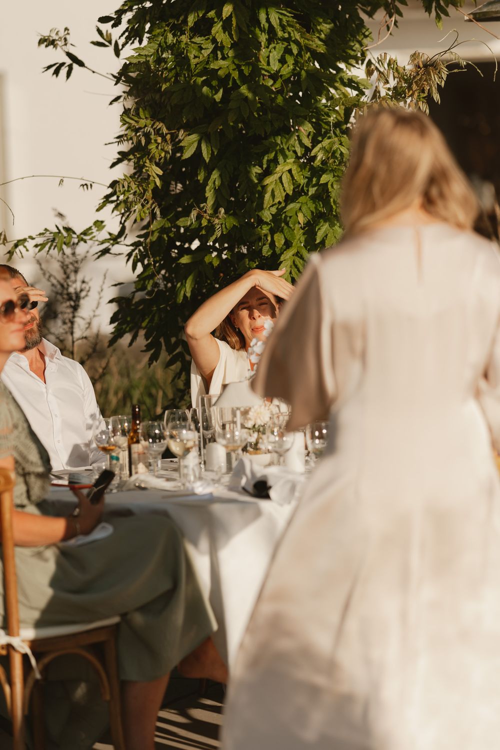 Guests seated at an elegant outdoor wedding reception table, bathed in warm golden hour sunlight with lush greenery.