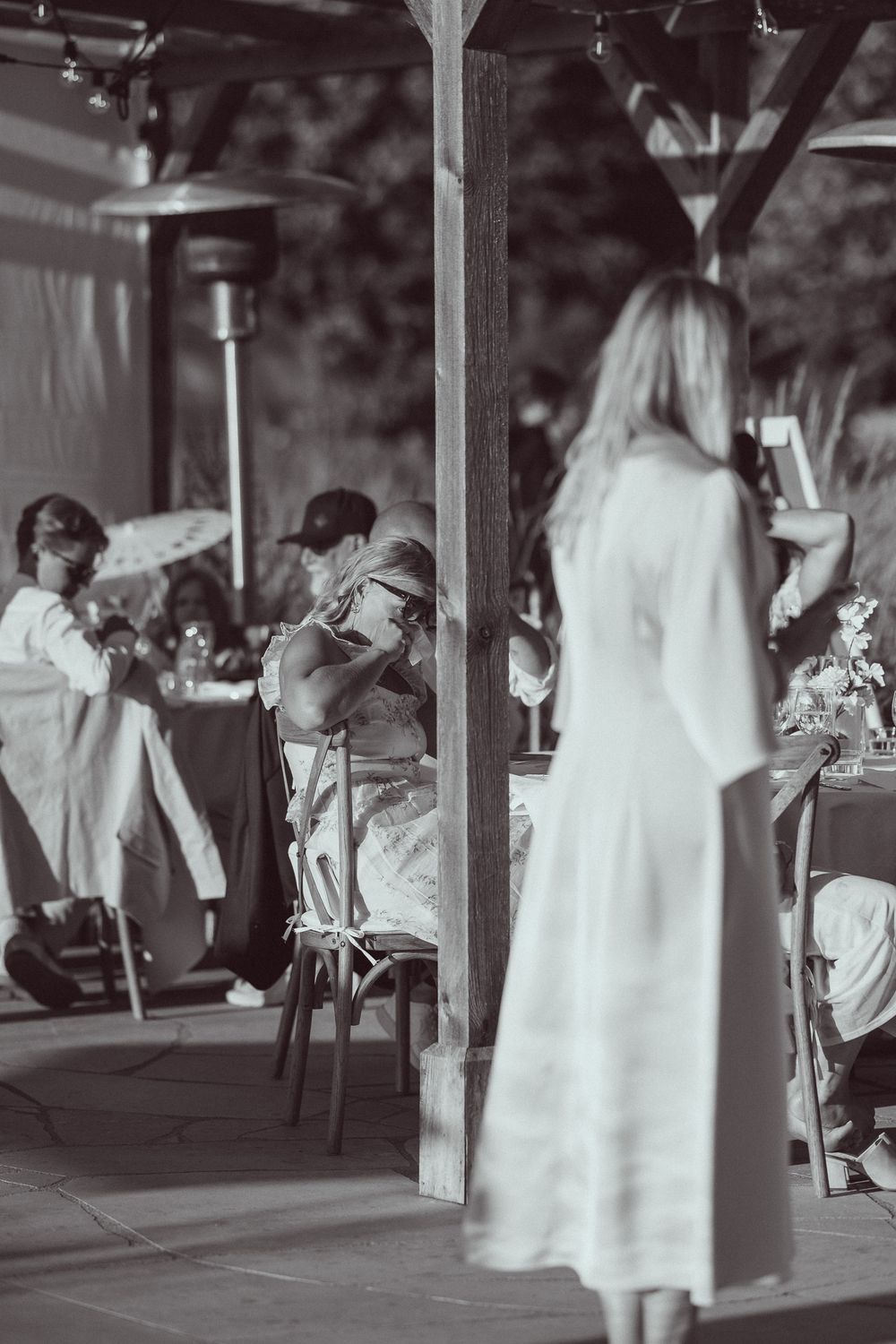 Black and white photo of guests at an outdoor wedding reception under a wooden pergola with string lights.