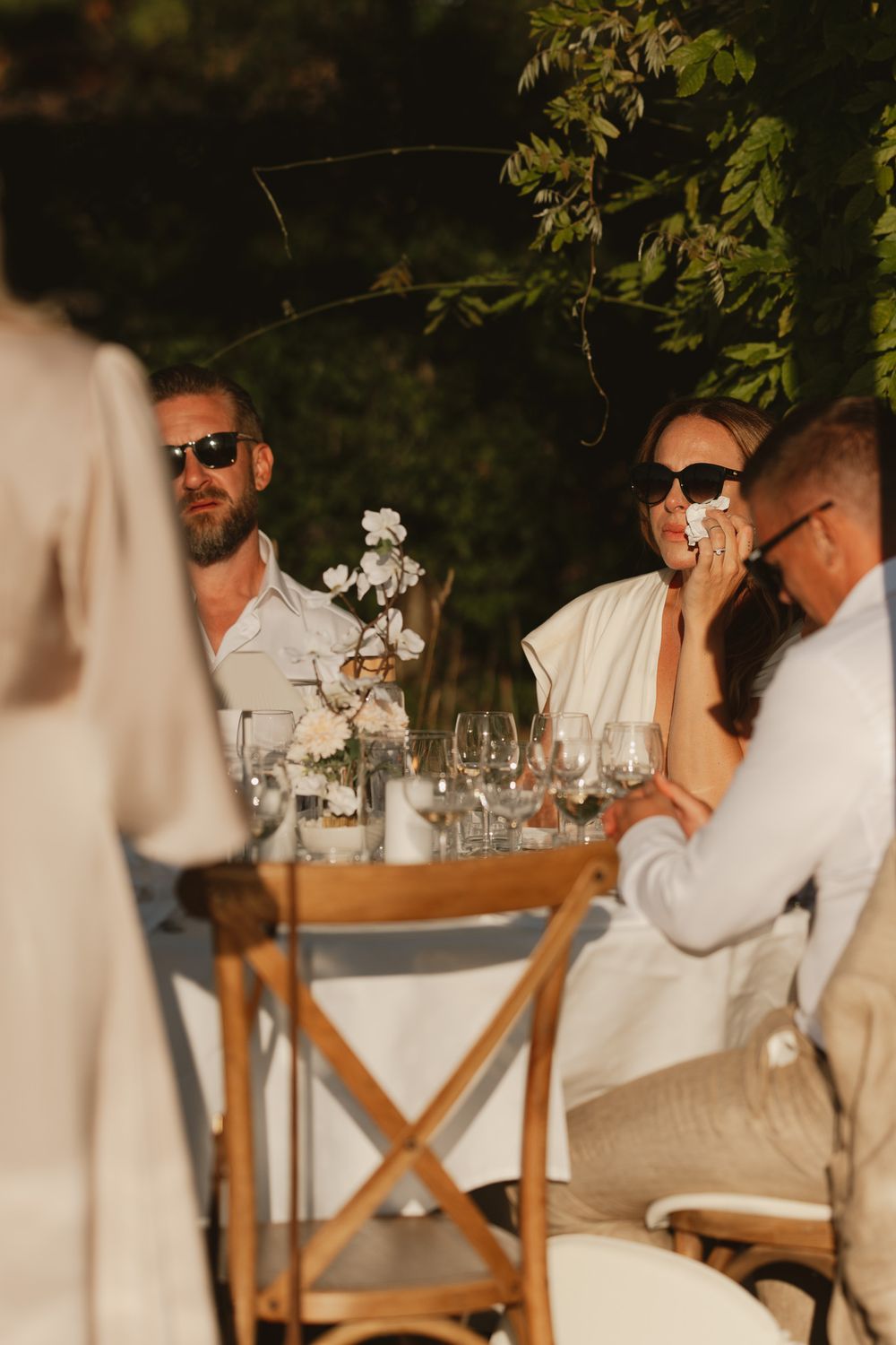 Guests in sunglasses enjoy an elegant outdoor dinner party at a white tablecloth table with floral centerpiece.