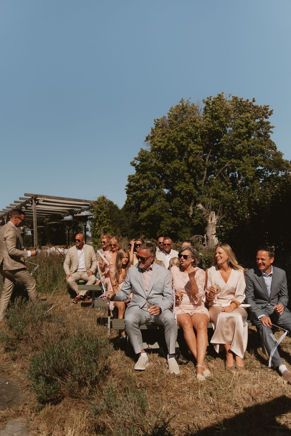 Guests seated outdoors at a sunny wedding ceremony, dressed in elegant attire, surrounded by lush green trees.