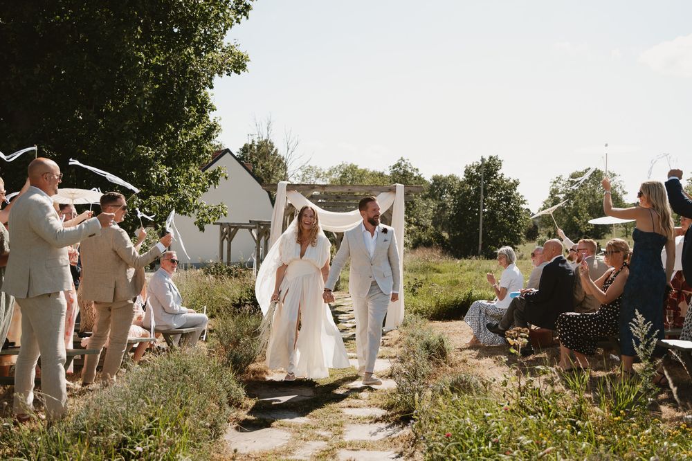 Bride and groom walk down outdoor wedding aisle surrounded by cheering guests on a sunny summer day.