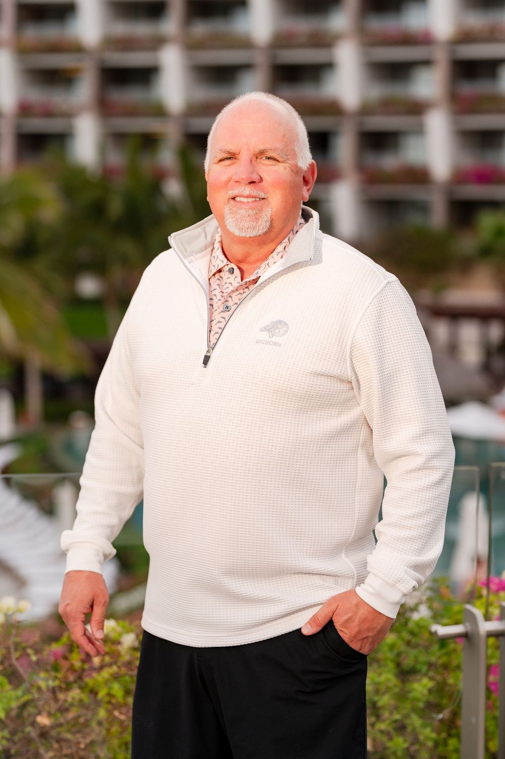 Man in white quarter-zip sweater smiling outdoors near tropical resort pool and gardens.