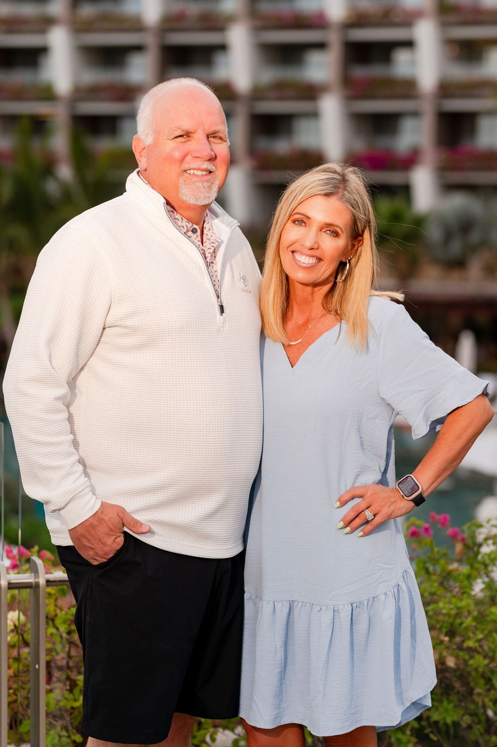 Smiling couple posing outdoors at a tropical resort with lush greenery and a hotel building in the background.