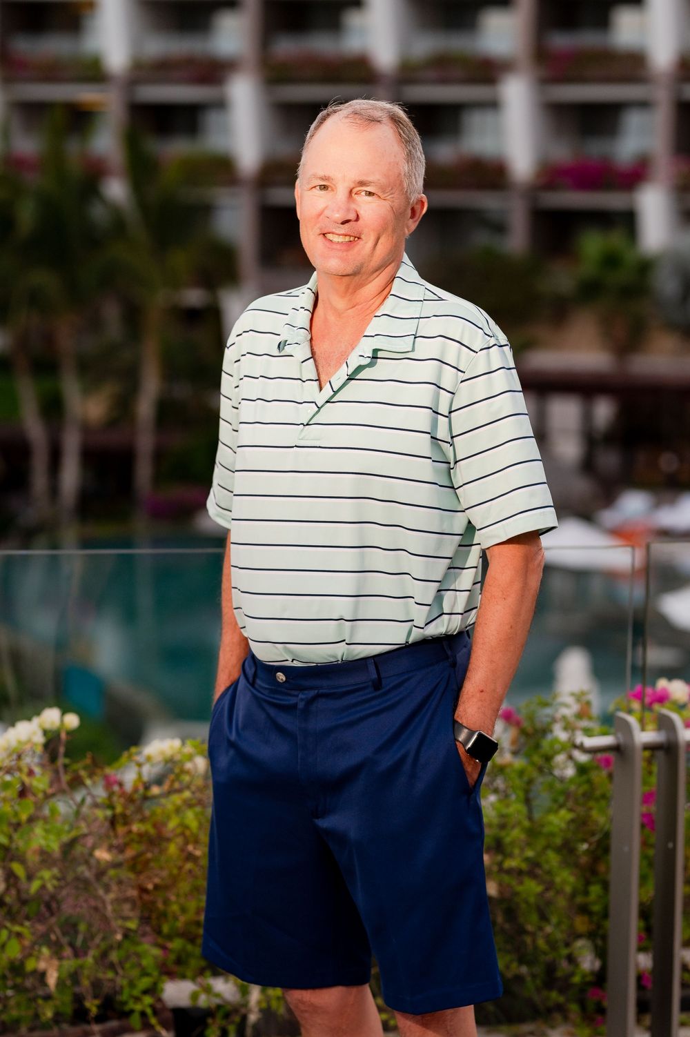Smiling middle-aged man in striped polo shirt and navy shorts standing by a resort pool with tropical landscaping.