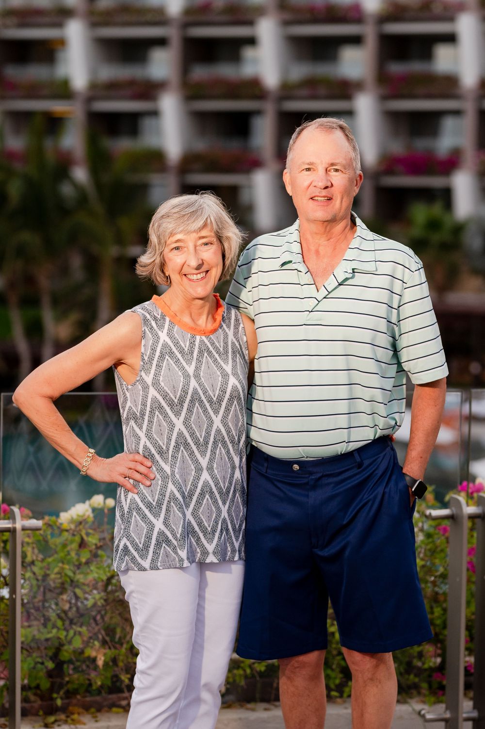Smiling senior couple posing together outdoors at a tropical resort with lush greenery and flowers in the background.