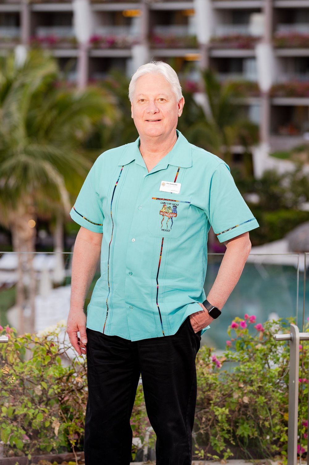 Man in teal uniform shirt with name tag stands confidently outdoors near a tropical resort pool and palm trees.