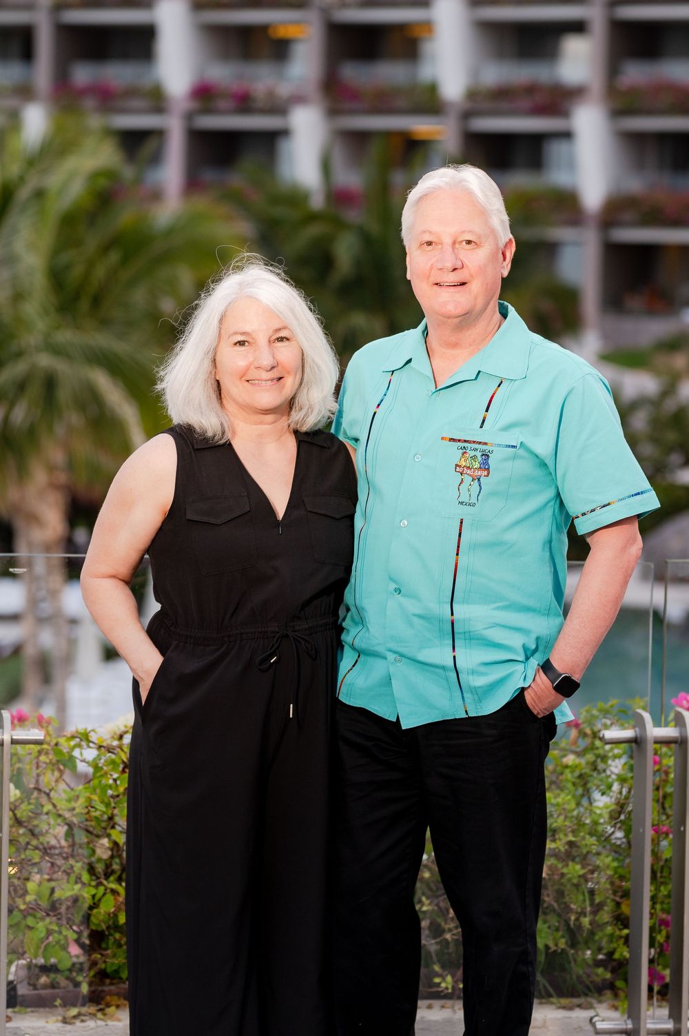 Smiling couple posing outdoors at a tropical resort, woman in black jumpsuit and man in teal shirt.