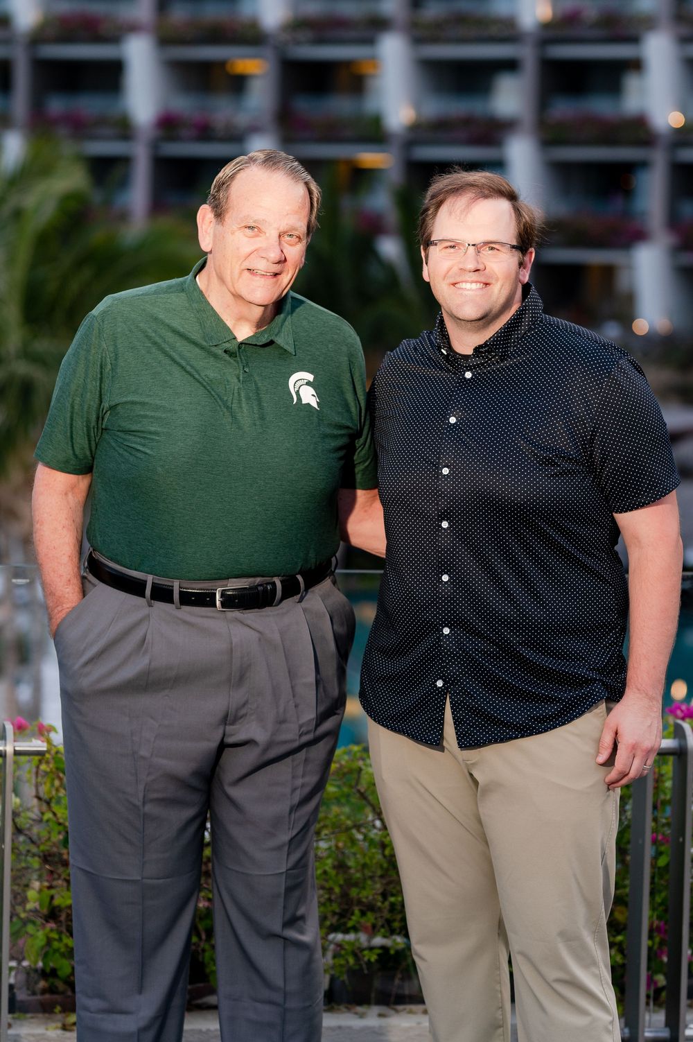 Two men smiling and posing together outdoors, one wearing a green Michigan State polo shirt, with a building in the background.