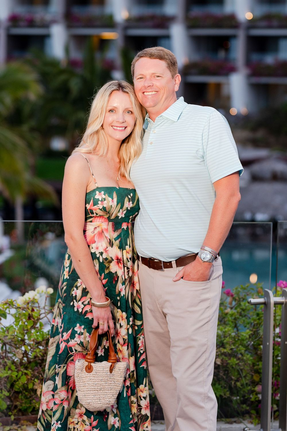 Couple posing together at a tropical resort, woman in floral dress, man in polo shirt, lush greenery background.