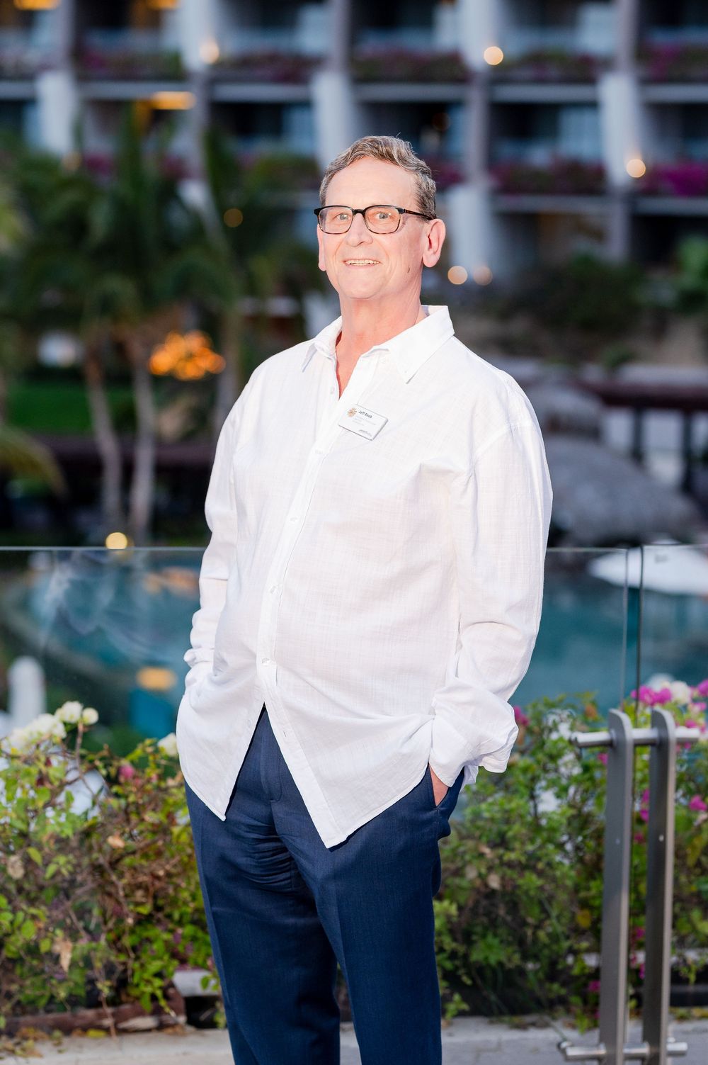 Man in white linen shirt and navy pants posing at a tropical resort with pool and hotel in background.