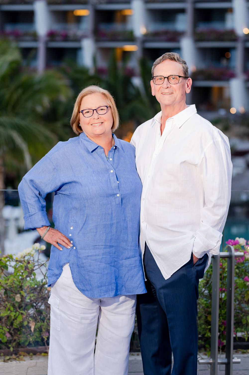A smiling couple in casual blue and white attire posing outdoors near a resort building with lush greenery.