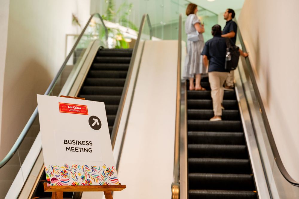 Business meeting sign on easel near escalator with attendees walking up in bright modern venue.