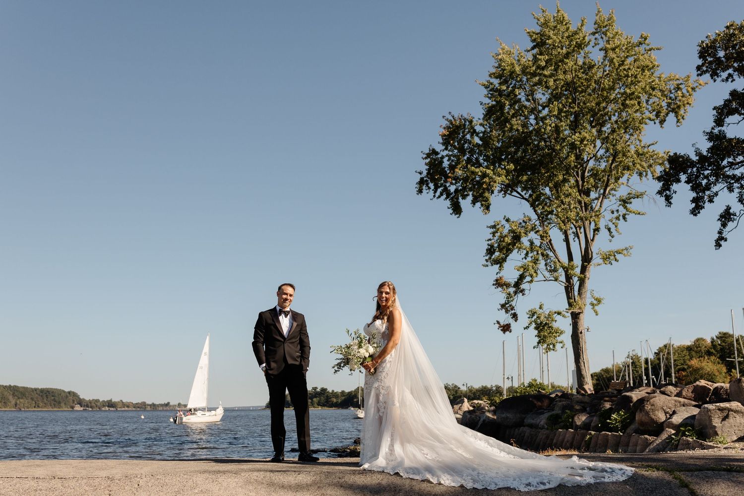 Bride and groom standing by a lakeside with a sailboat and trees in the background on a sunny wedding day.