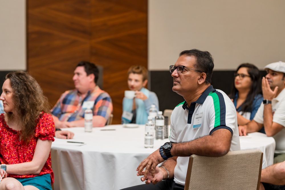 Diverse group of adults seated at round tables in a conference room, attentively listening during a seminar or meeting.