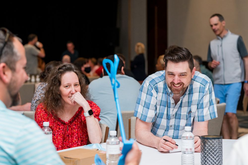 Two smiling adults sit together at a table during a casual indoor event, engaged in friendly conversation.