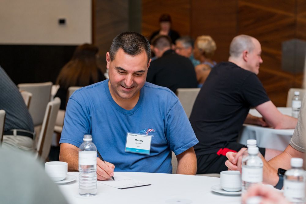 Man in blue t-shirt smiling while writing notes at a round table during a professional networking event.