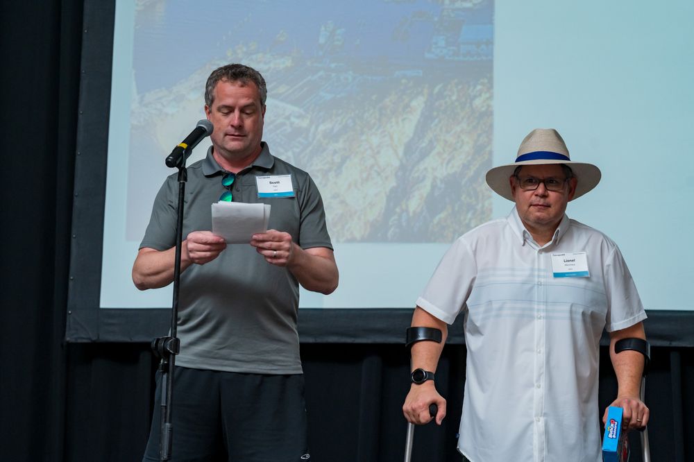 Two men on stage presenting, one reading papers at microphone, other standing with crutch before projected mountain image.