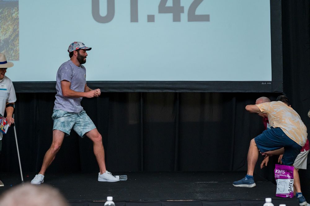 Two men compete in a timed challenge on stage, one lunging forward while the other bends down, with 01:42 displayed behind them.