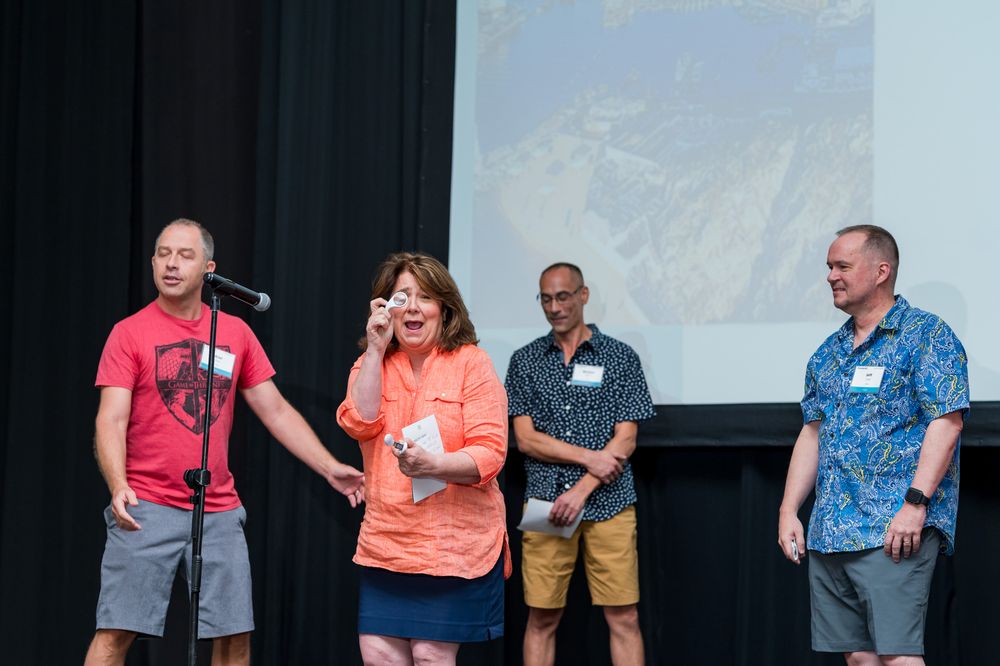 Four adults stand on stage at an event, with a woman in orange speaking into a microphone while others smile beside her.