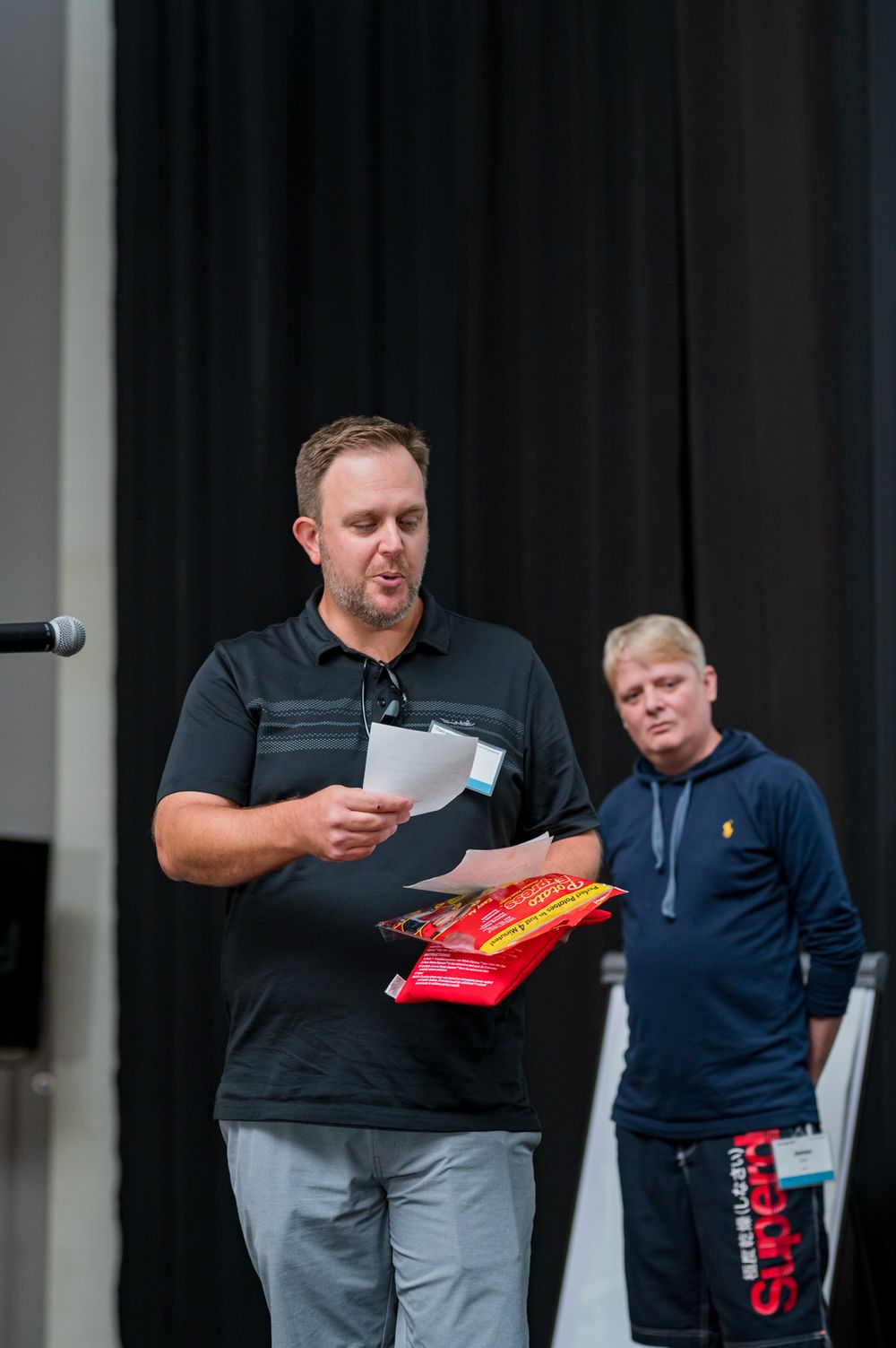 Man in black polo shirt reads paper while holding snack bag, second man in blue Superdry hoodie stands behind him.