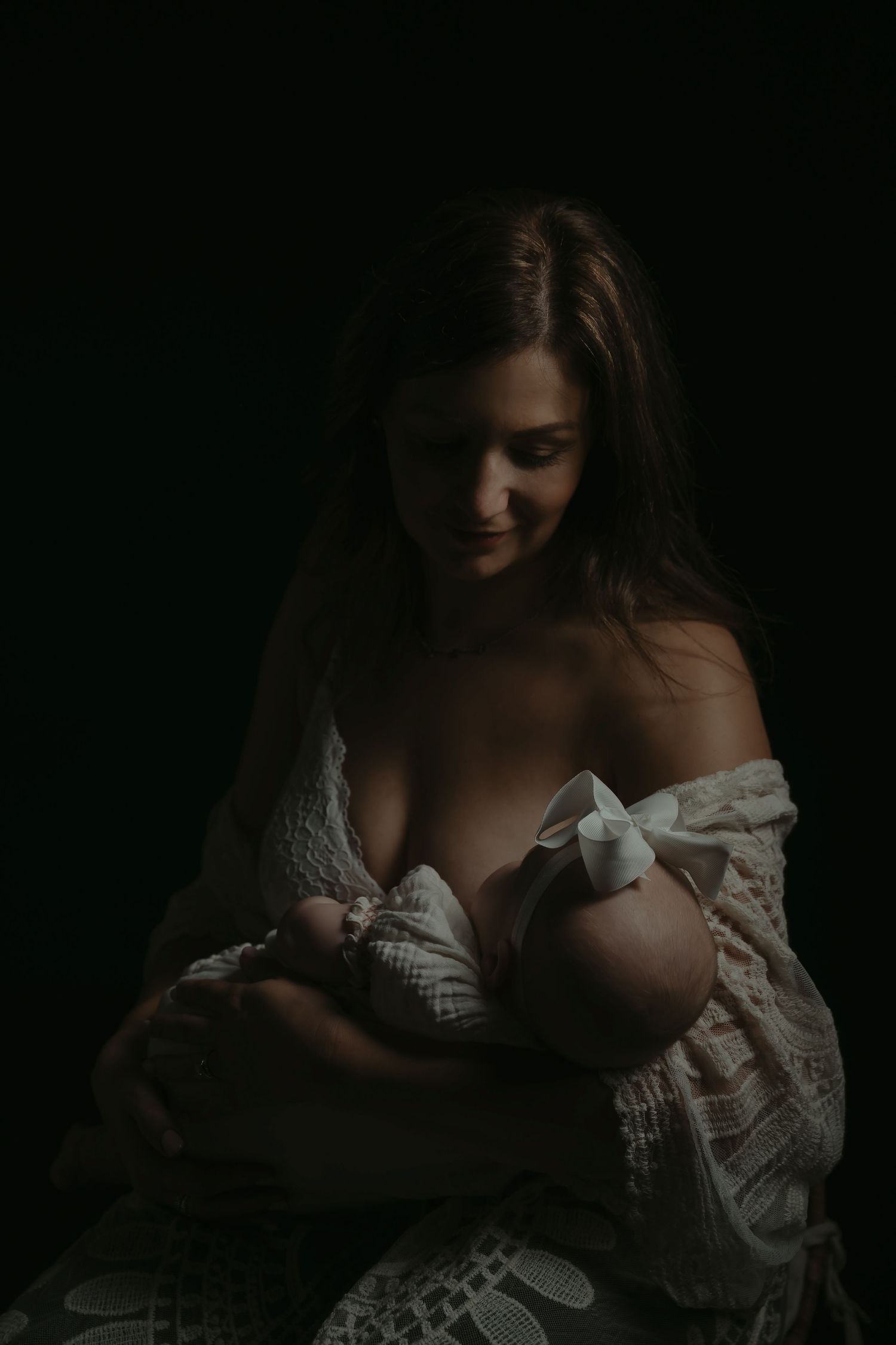 A mother tenderly breastfeeds her newborn baby in a dark, moody portrait with soft, dramatic lighting.