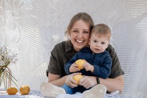 A smiling mother holds her baby boy who clutches a lemon, sitting against a white lace backdrop with flowers nearby.