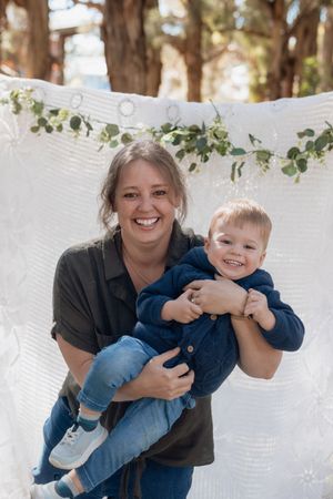 A smiling mum holds a happy toddler boy in blue outfit outdoors with white floral backdrop and trees in a mother and child photoshoot.