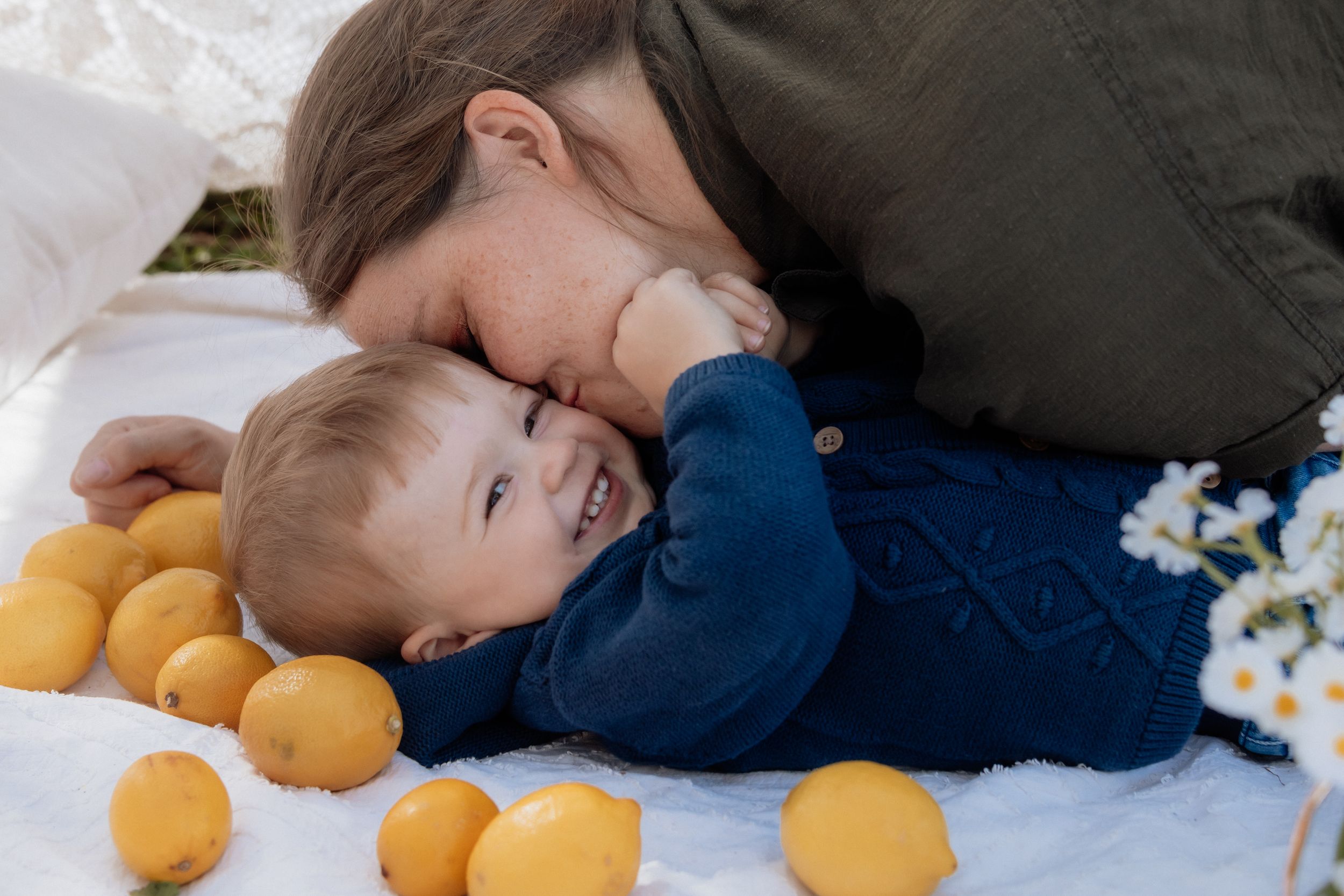 Mother lovingly nuzzles smiling baby in blue outfit on white blanket surrounded by fresh lemons and daisies in a park in Rouse Hill.