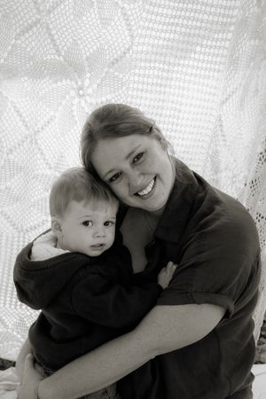 Black and white photo of a smiling young mother hugging her baby in front of a lace curtain backdrop.